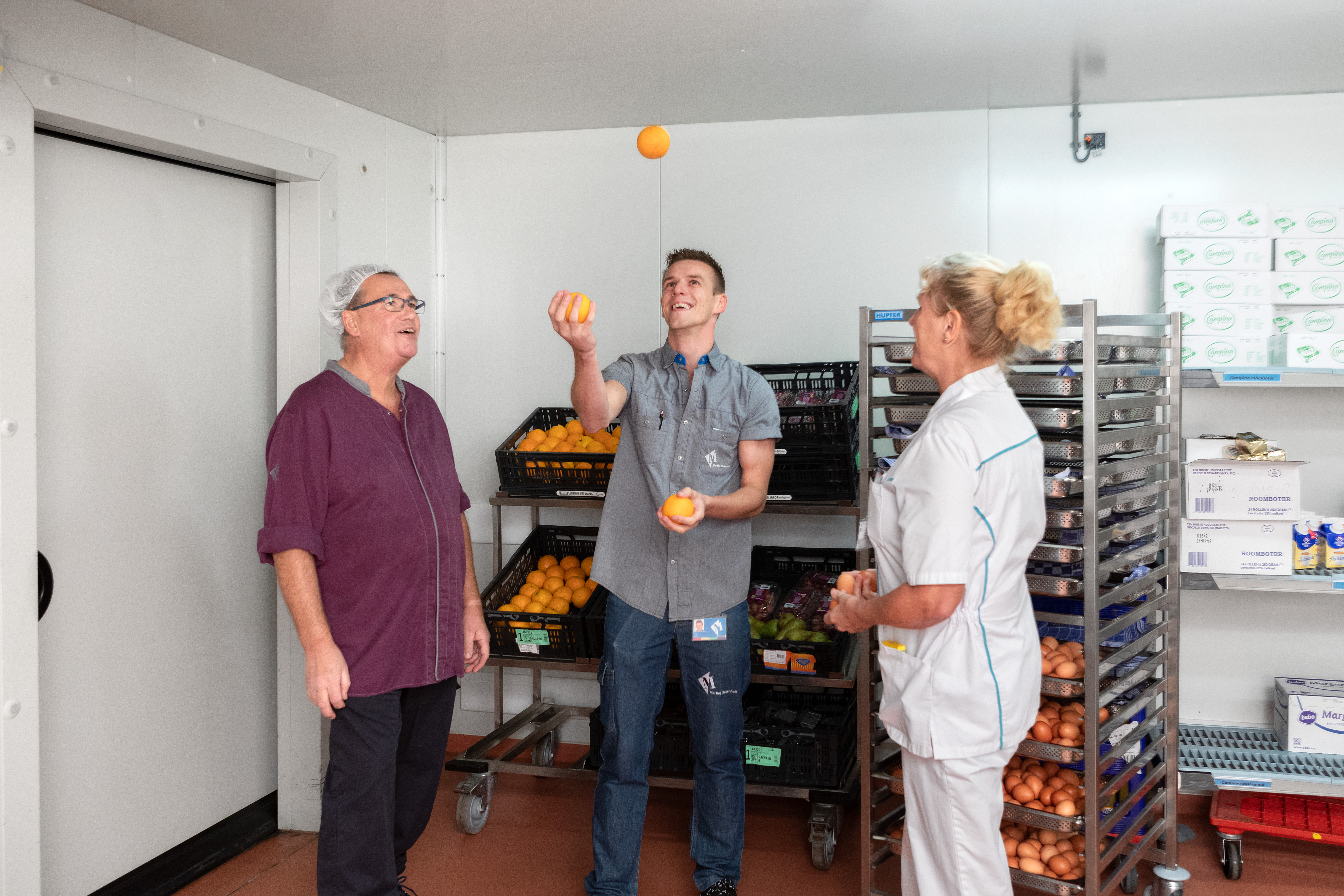 Een medewerker jongleert met sinaasappels. Twee medewerkers kijken toe. 